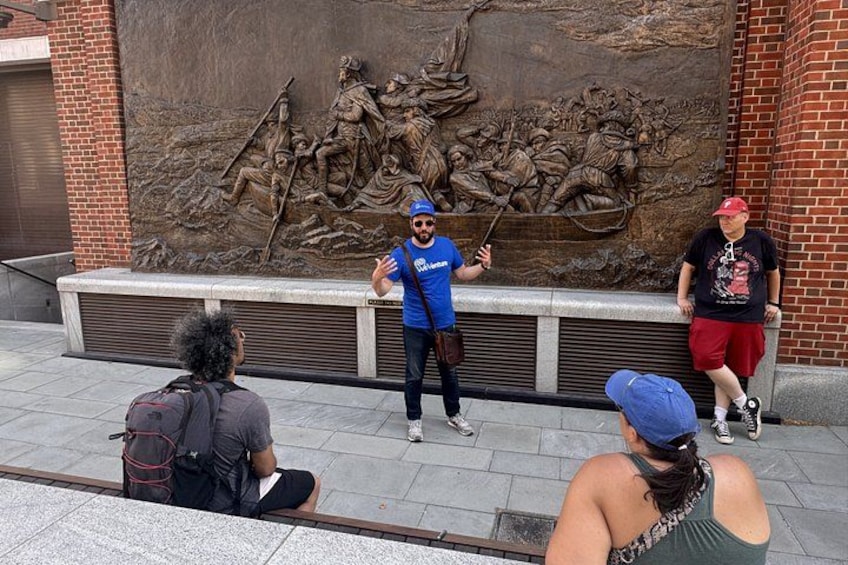 Une sculpture en relief en bronze ornant le mur d’un bâtiment, une personne faisant un geste, et trois personnes assises sur le trottoir. 4
