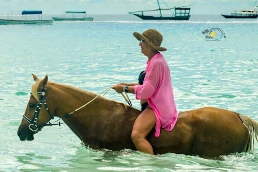 Eine Person reitet auf einem Pferd durch flaches Wasser, im Hintergrund sind Boote zu sehen. 3