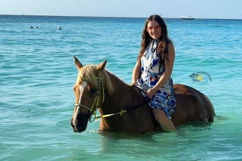 Eine Person reitet auf einem Pferd in klarem türkisblauem Wasser, im Hintergrund ein Boot. 1
