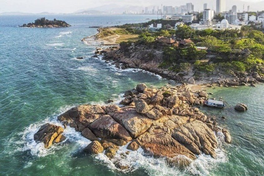 A rocky coastline with waves crashing against the rocks, a small island in the distance, and a city skyline in the background. 4