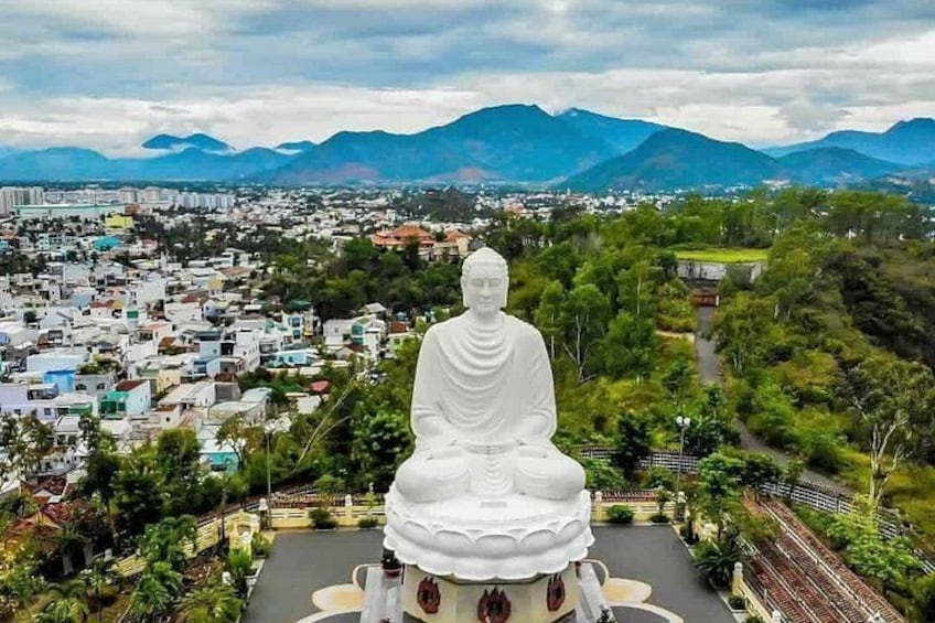 A large white statue of a seated figure with a lotus base, surrounded by greenery, overlooking a cityscape with mountains in the background. 3