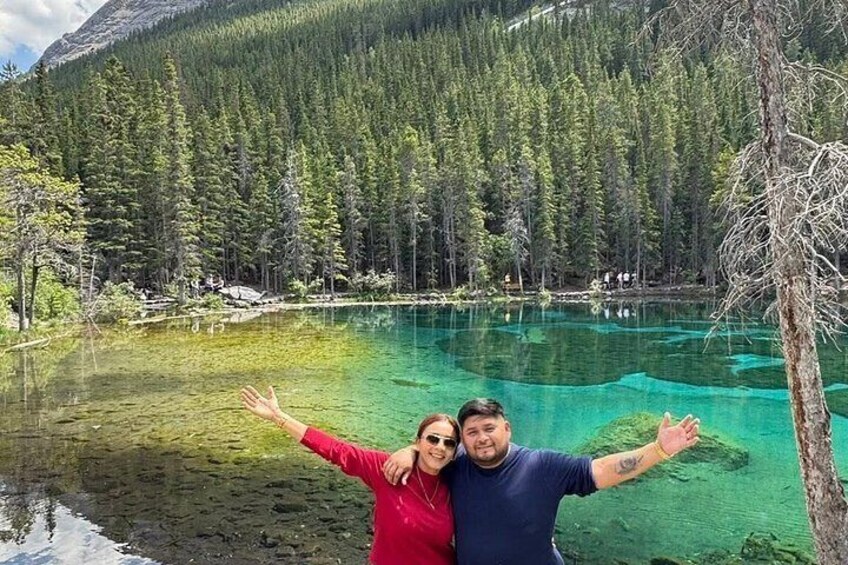 A couple standing by a clear turquoise lake surrounded by dense forest and mountains. 2