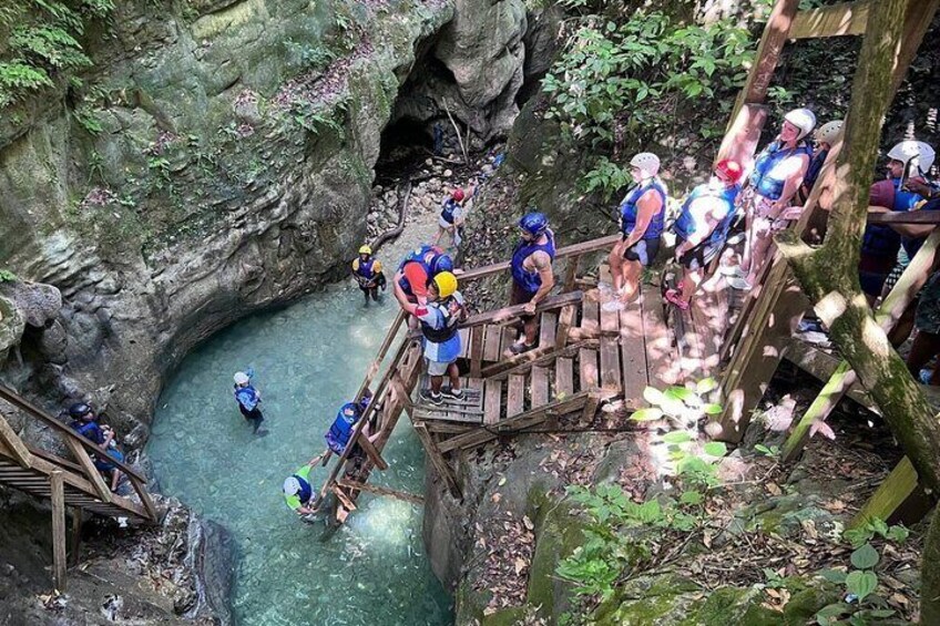 A group of people in blue life vests and helmets on a wooden bridge over a river in a rocky canyon. 5