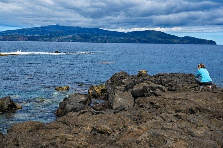 A rocky shoreline with a person sitting on the rocks, a body of water, and mountains in the distance. 3