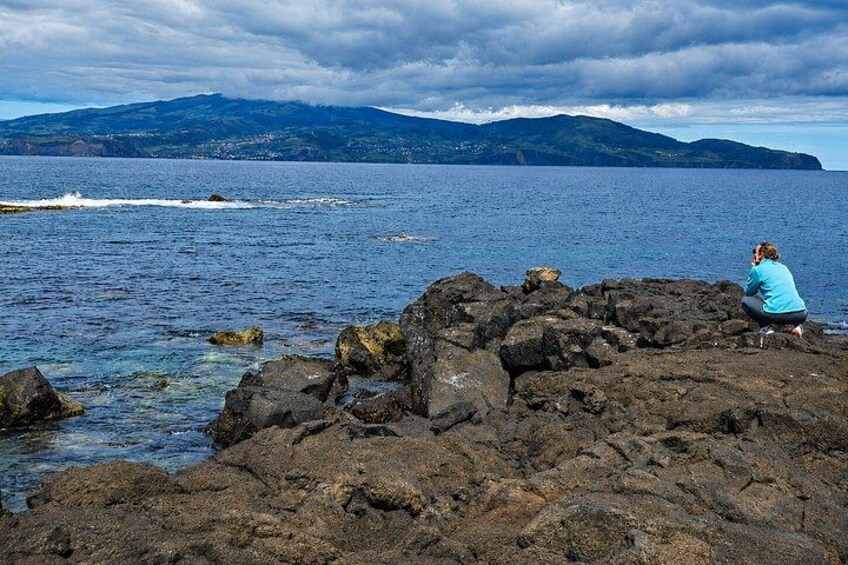 A rocky shoreline with a person sitting on the rocks, a body of water, and distant mountains under a cloudy sky. 3
