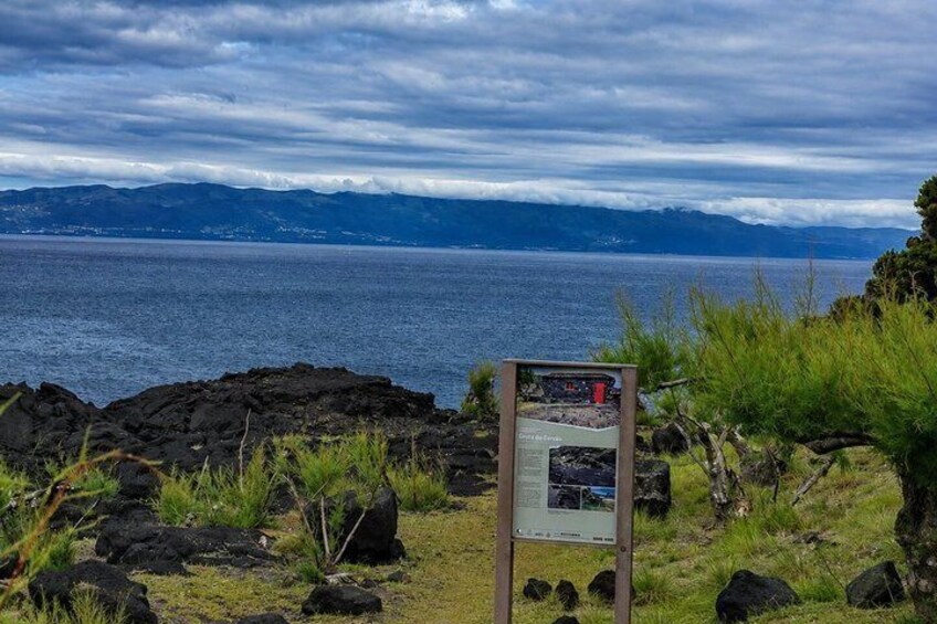 A coastal landscape with a rocky foreground, a signboard, and a view of the ocean and distant mountains under a cloudy sky. 4