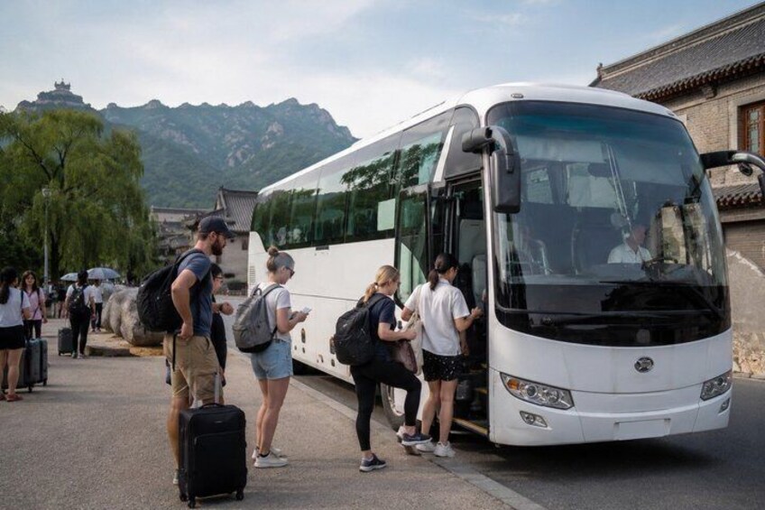 A group of people boarding a white bus with luggage, near a traditional building with a mountain in the background. 2