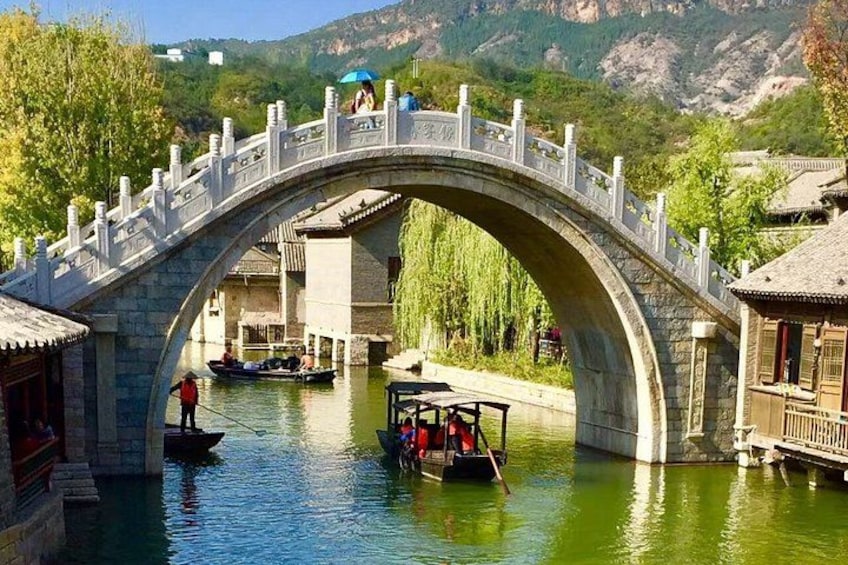 A stone bridge over a river with boats, traditional buildings, and a mountainous backdrop. 2