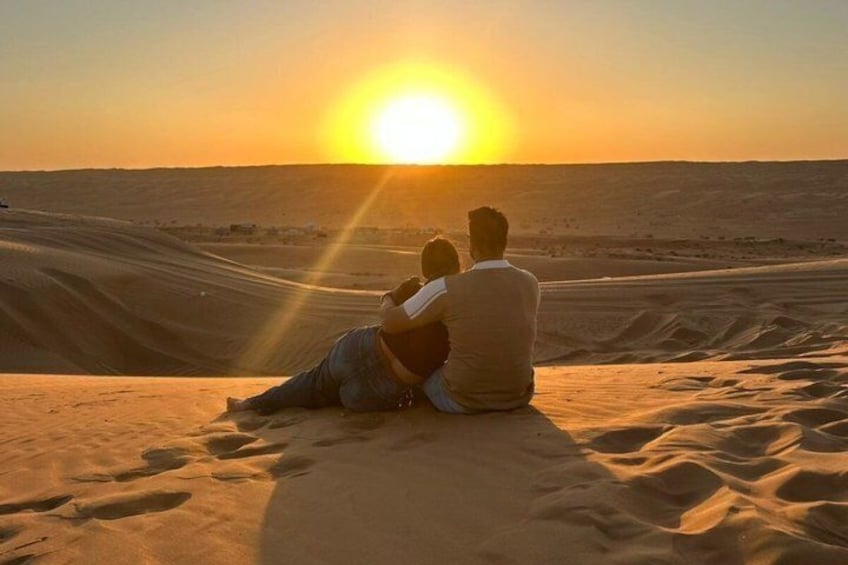 Two people sitting on a sand dune, facing the sunset over a desert landscape. 1