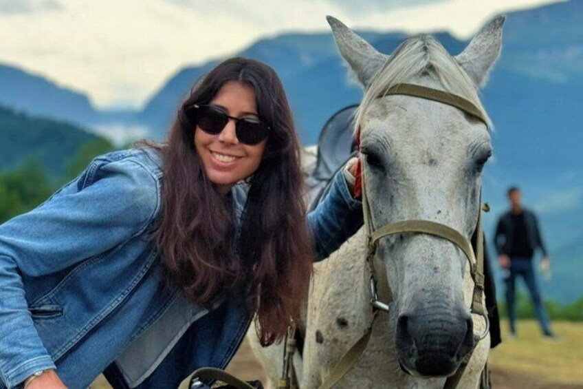 A person in a denim jacket petting a white horse with a mountainous landscape in the background. 2