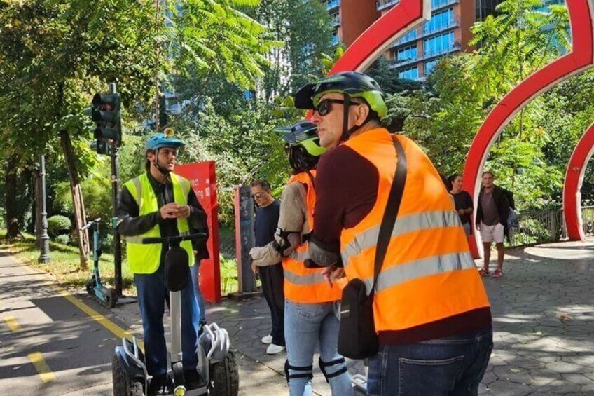 People in safety vests standing on a sidewalk near a traffic light and a red sculpture. 4