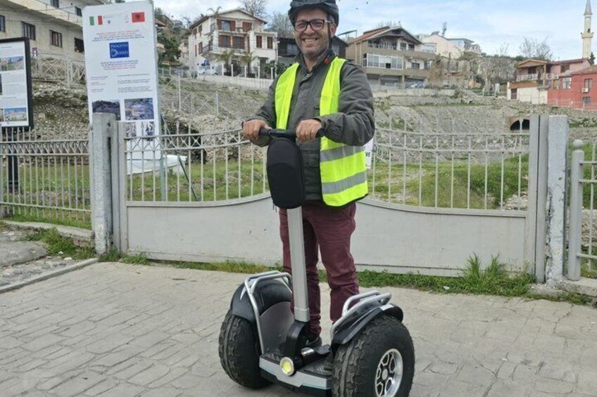 A person on a Segway, wearing a safety vest, in an urban setting with buildings and a fence in the background. 3
