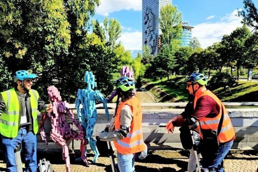 A group of people in safety vests and helmets standing on a sidewalk, with trees and a building in the background. 1