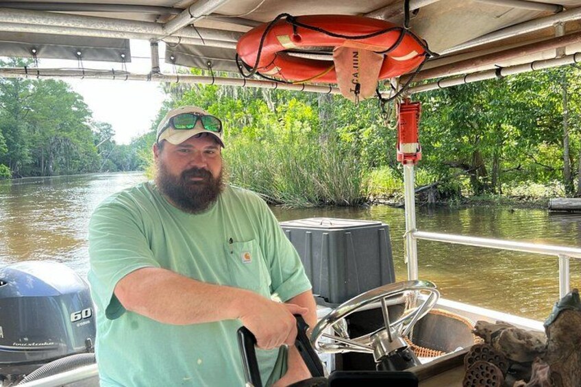 A man steering a boat on a river, surrounded by greenery and a life preserver hanging from the boat's structure. 4