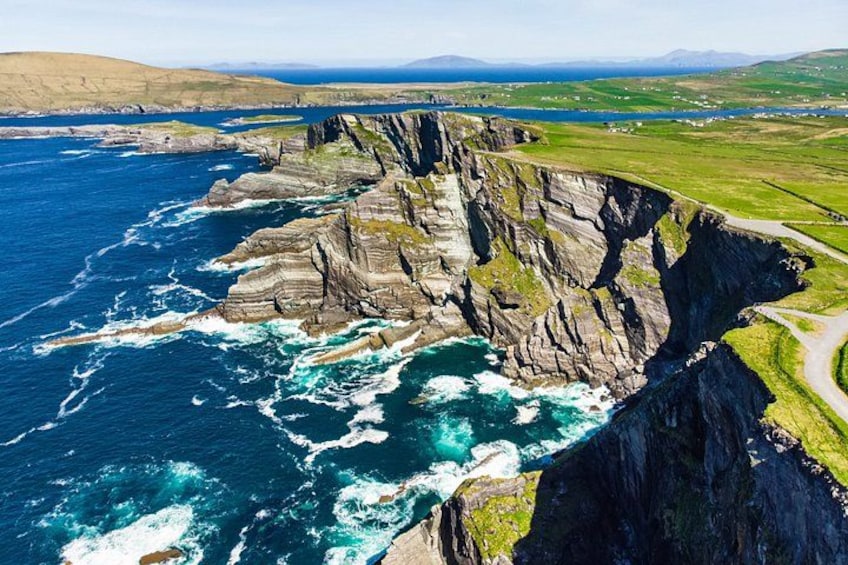 A coastal cliff with waves crashing against it, a road along the cliff, and a distant view of the sea and hills. 1