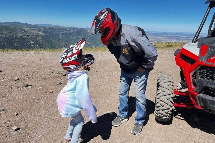 A person wearing a helmet and a jacket standing next to a red ATV on a dirt path with a mountainous landscape in the background. 5