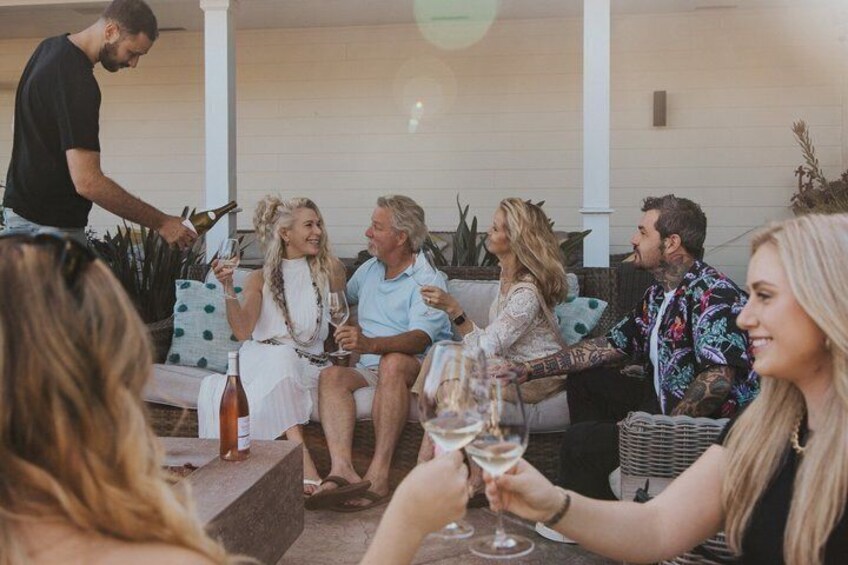 A group of people sitting on outdoor furniture, holding glasses of wine, with a person pouring drinks. 1