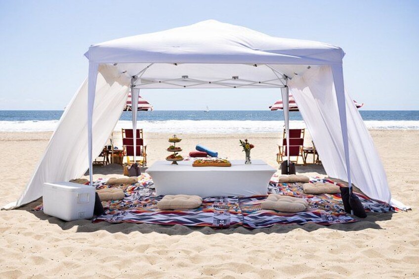 A beach tent with a table and chairs, a blanket, and a small stage with a statue. 2
