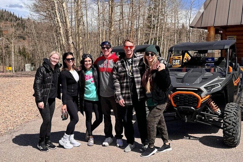A group of people posing with a large off-road vehicle in a wooded area. 1