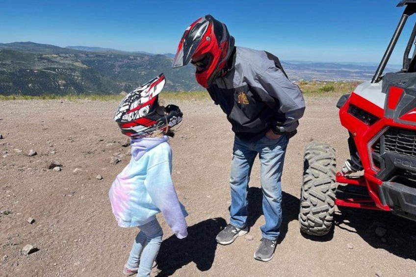 A person wearing a helmet and a jacket standing next to a red ATV on a dirt path with a mountainous landscape in the background. 5