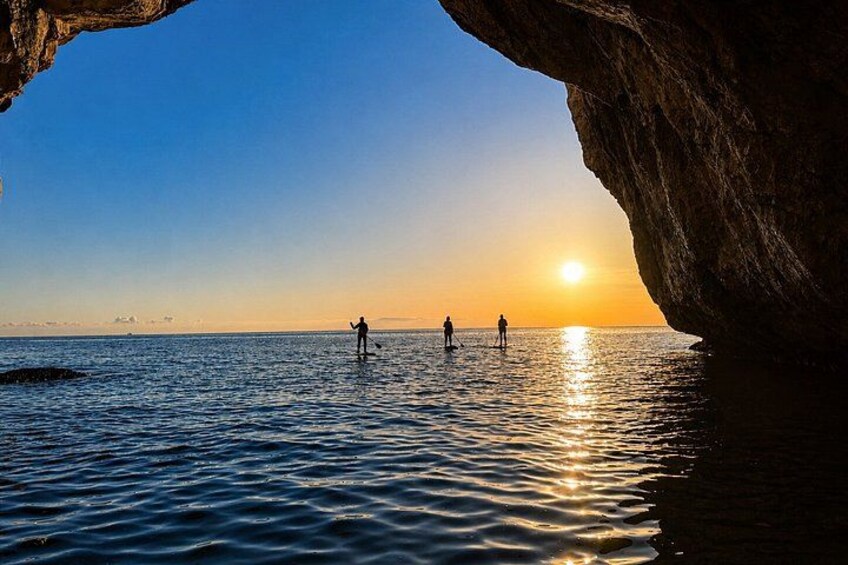 A sunset over water with a cave opening, people standing in the water, and a silhouette of a person. 3