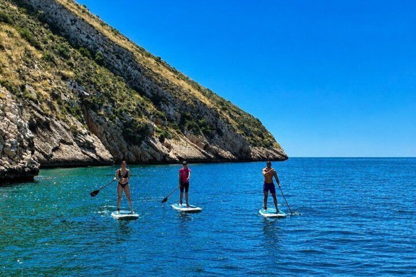Three people paddleboarding on a calm sea with a rocky coastline in the background. 1