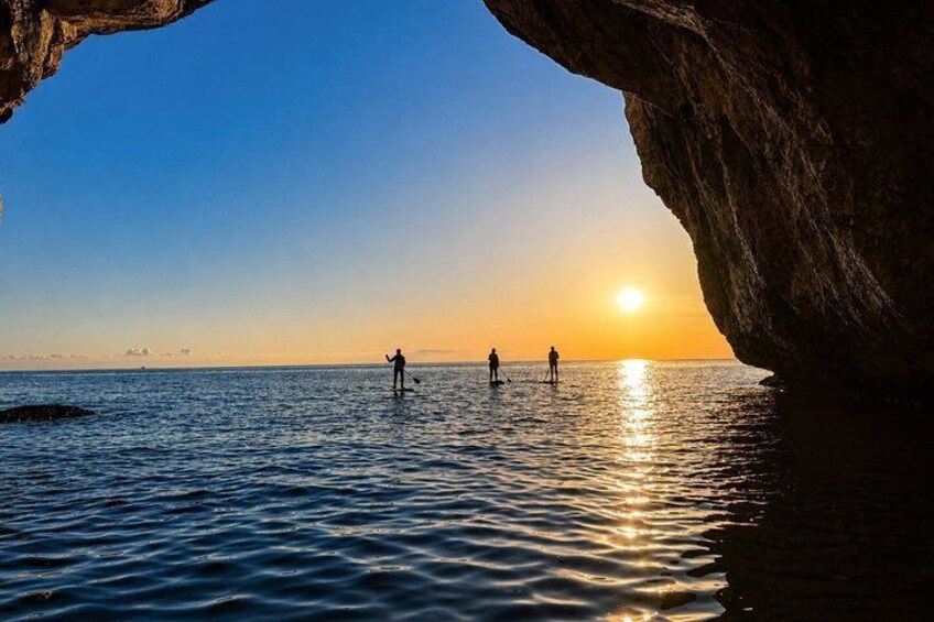 A sunset over a calm sea with people paddleboarding near a rocky cave entrance. 3
