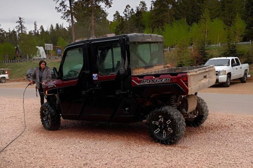 A red DANGER off-road vehicle with a man using a winch on a gravel surface, surrounded by trees and a white pickup truck in the background. 3