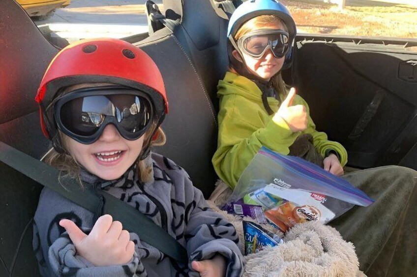 Two children in a car, wearing helmets and seatbelts, with a book and snacks visible. 4