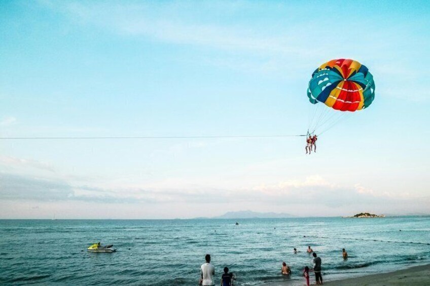 Dos personas haciendo parasailing sobre una playa, con gente nadando en el agua. 3