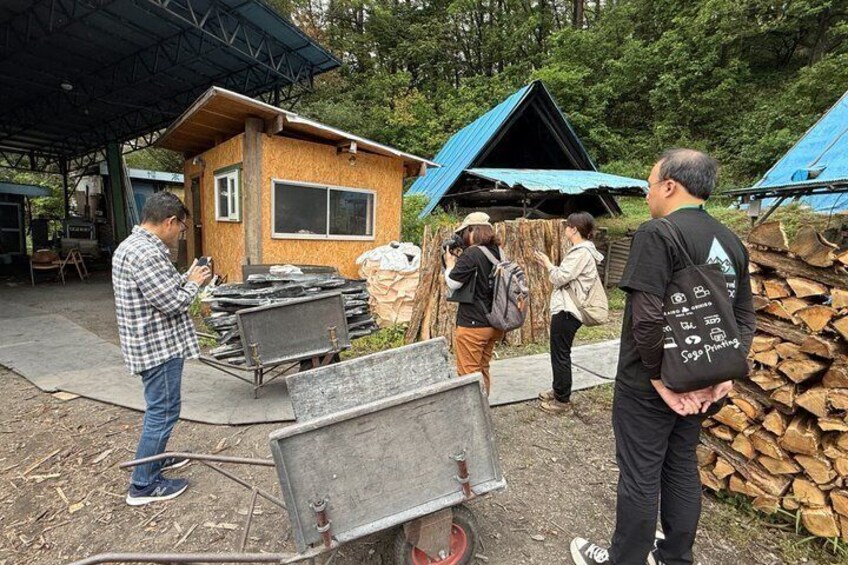 A group of people near a small wooden structure with a blue roof, a wheelbarrow, and a stack of firewood. 3