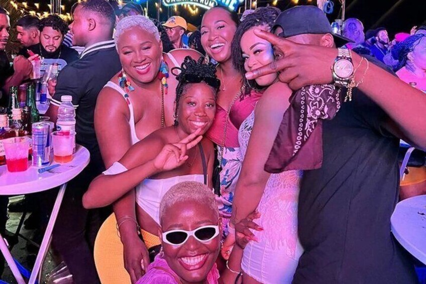 A group of people at an outdoor event with tables, drinks, and a neon sign in the background. 5
