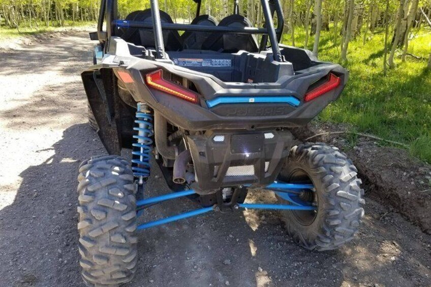A Polaris RZR off-road vehicle parked on a dirt path with large tires and blue suspension components. 2