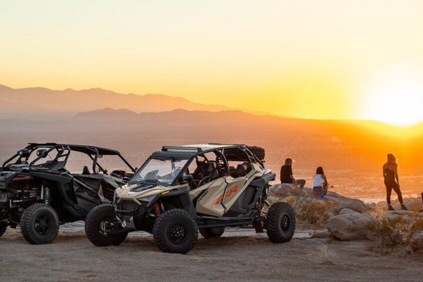 Two off-road vehicles parked on a rocky shore with a sunset in the background. 1