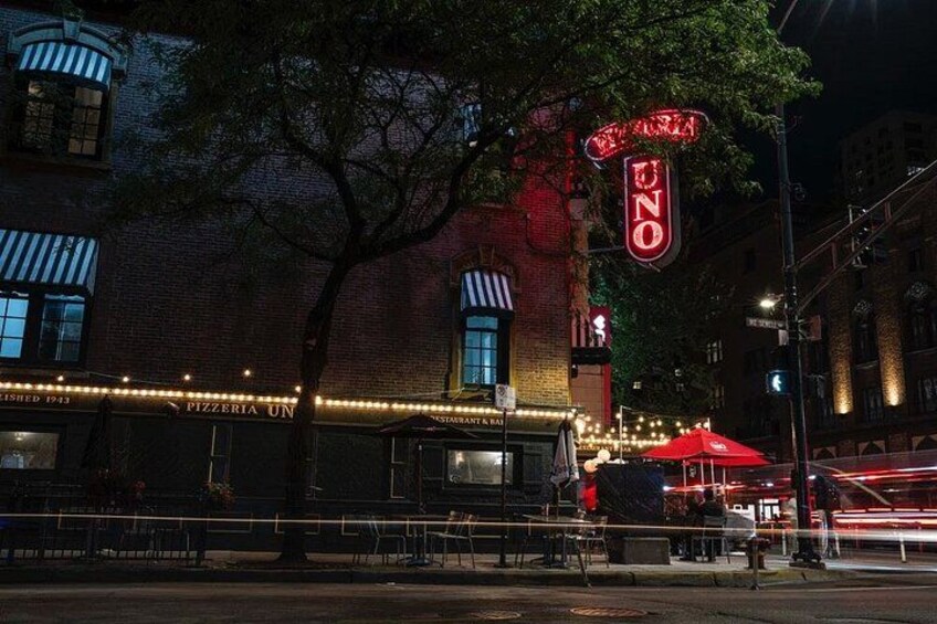 A brick building with a neon sign that reads 'UNO PIZZERIA UN' and a red umbrella. 2