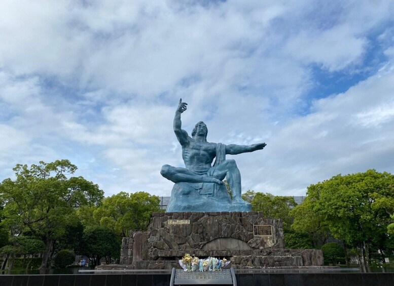 A large bronze statue of a seated man with one arm raised, set against a backdrop of trees and a cloudy sky. 1