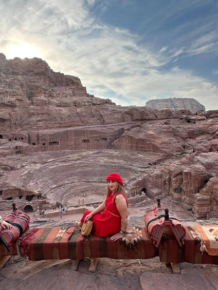 A person in a red dress sitting on a bench with a large amphitheater in the background. 2