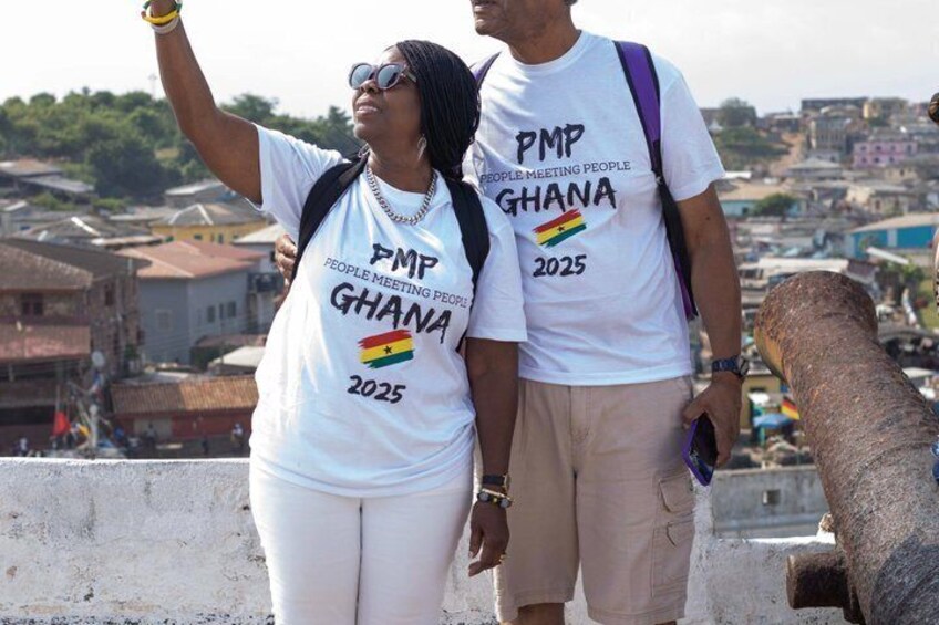 Two people wearing white t-shirts with 'PMP Ghana 2025' printed on them, standing on a rooftop with a cityscape in the background. 2