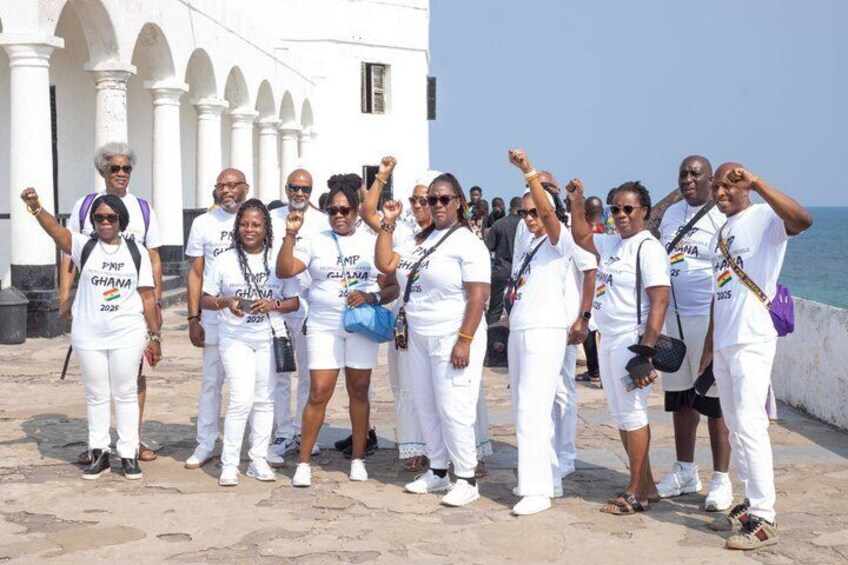 A group of people in white attire with 'GHANA 2022' on their shirts, standing on a paved area with a white building and a body of water in the background. 4