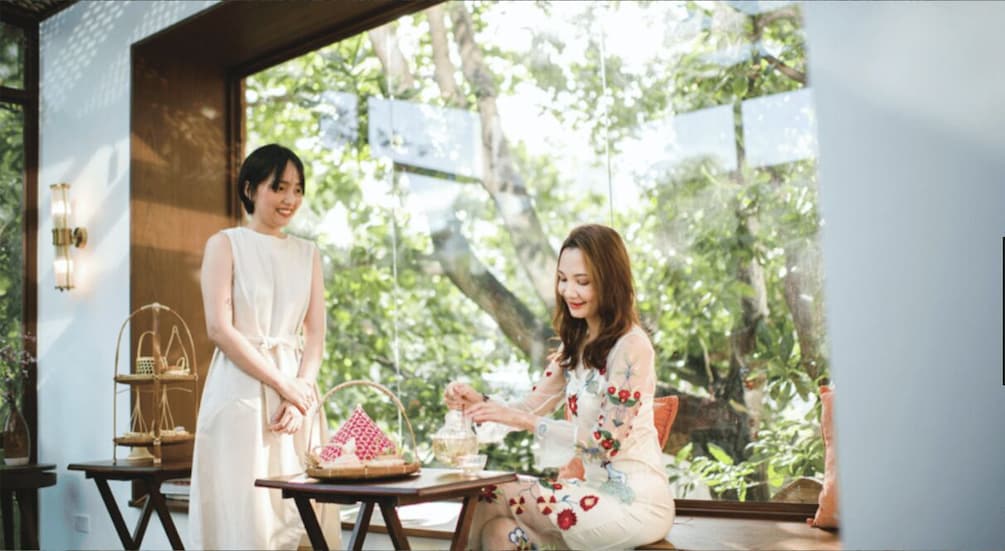 Two people sitting at a table with a basket and teapot, in a room with a large window overlooking greenery. 4