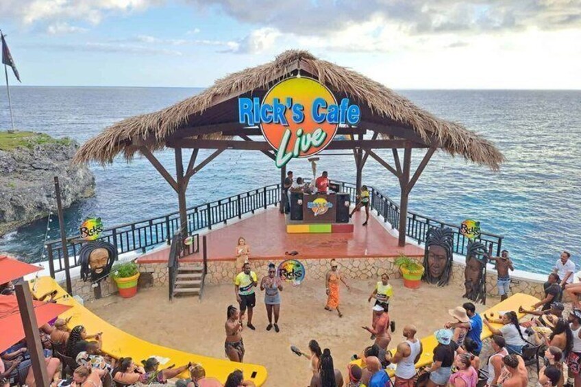 A beachside stage with a thatched roof labeled 'Rick's Cafe Live' and a crowd of people. 5