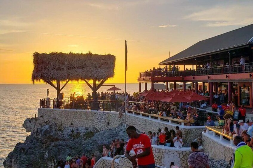 A sunset at a coastal restaurant with a thatched structure and outdoor seating. 1