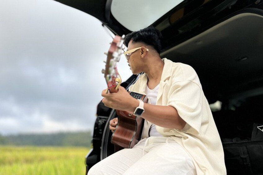 A person playing a guitar in a car with a rural landscape in the background. 1