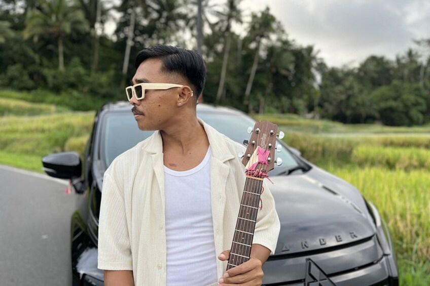 A person holding a guitar standing in front of a car with a rural landscape in the background. 2