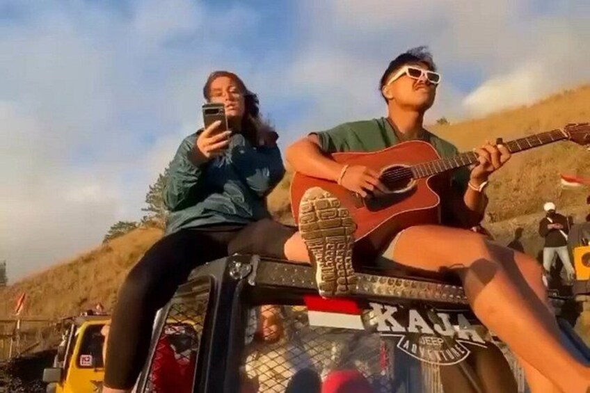 Two people sitting on a vehicle with a guitar, a person taking a photo, and a vehicle with a logo. 3