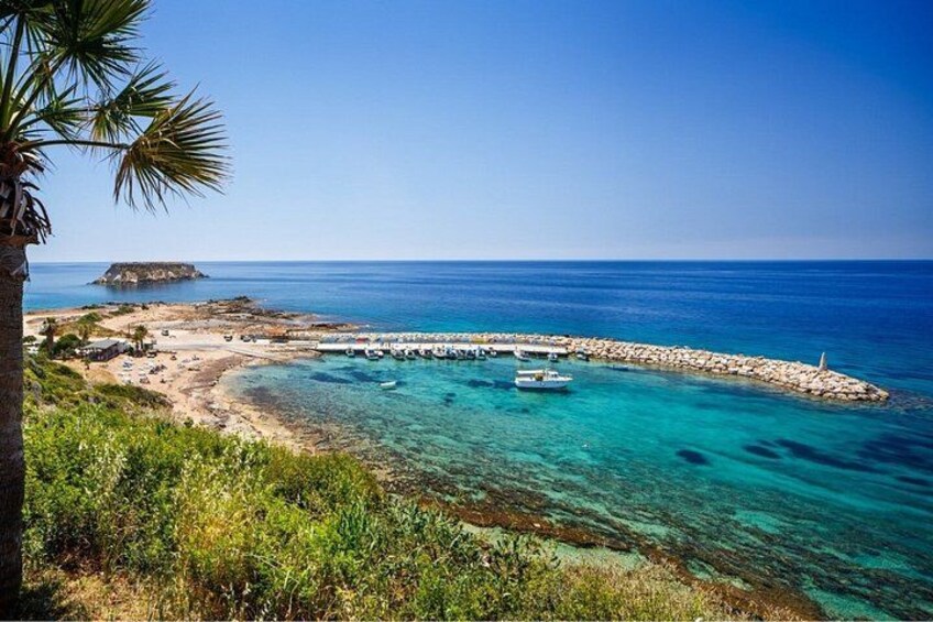 A coastal landscape with clear turquoise waters, a sandy beach, a rocky breakwater, and a small island in the distance. 3