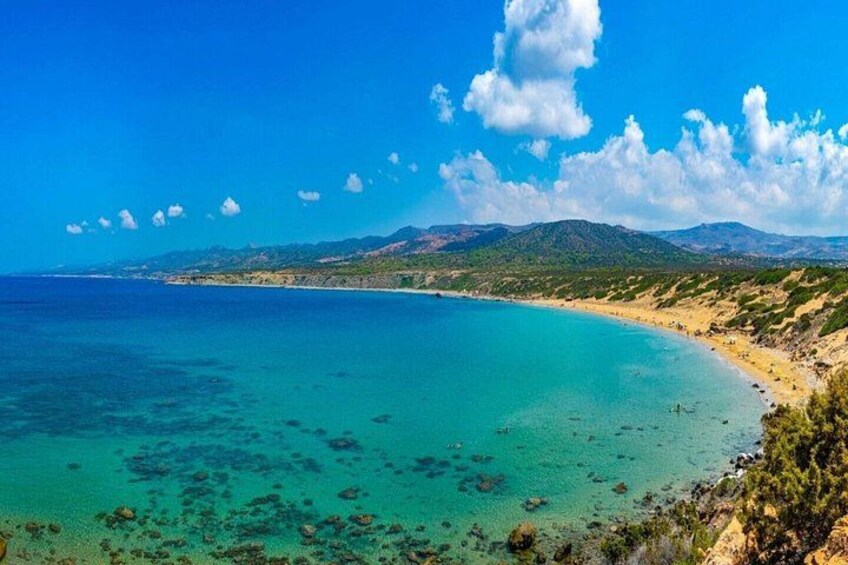 A coastal landscape with clear turquoise waters, sandy beaches, and distant mountains under a blue sky with scattered clouds. 5