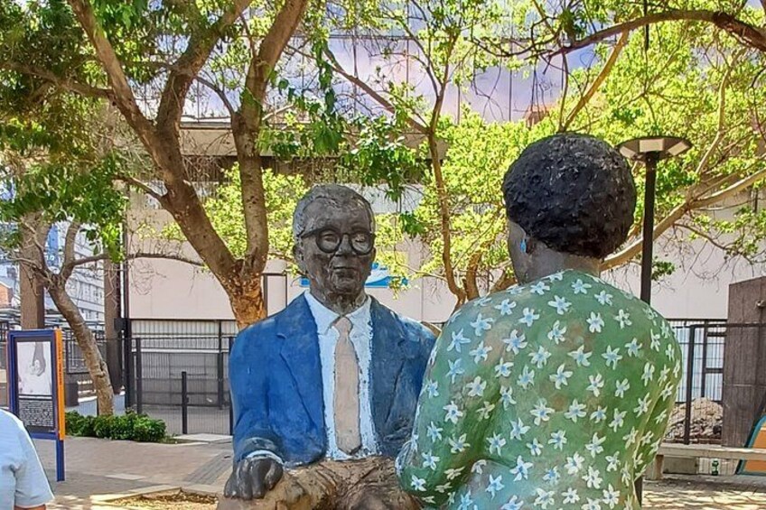 A statue of a man in a suit and tie, a tree, and a person in a green floral shirt. 1
