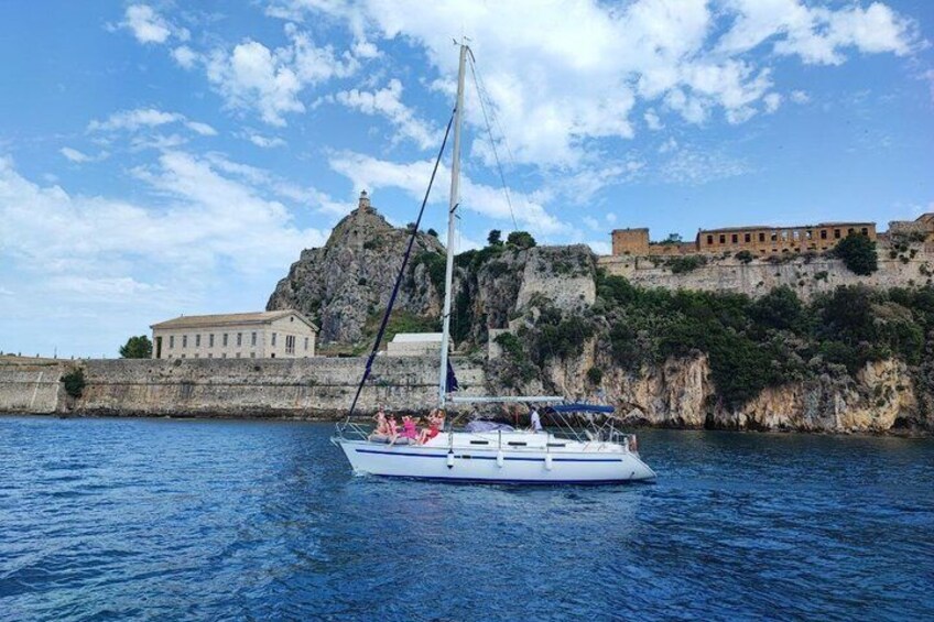 A sailboat on calm waters with a historic building and fortifications in the background. 1