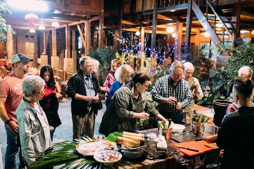 A group of people gathered around a table with food and drinks, in an indoor setting with wooden beams and plants. 1